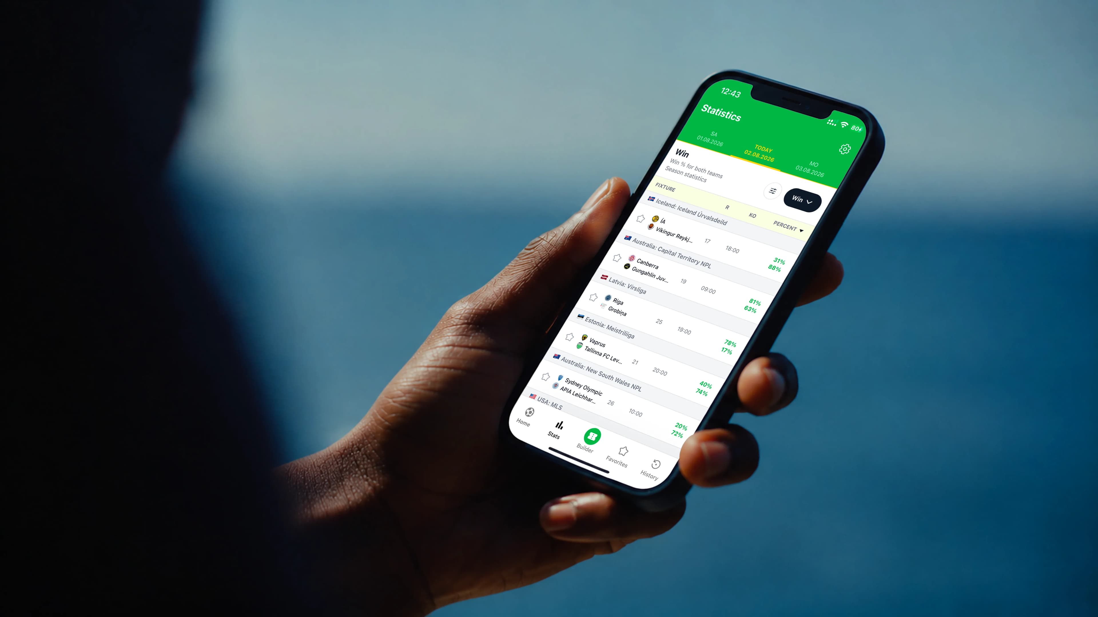 Hand holding an iPhone showing the Winsday football statistics app against a coastal blue backdrop